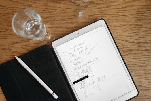 Top-down view of a tablet with handwritten notes, stylus, and glass on a wooden surface.