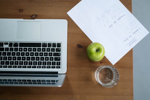 Top view of a workspace with a laptop, green apple, water glass, and paper notes.